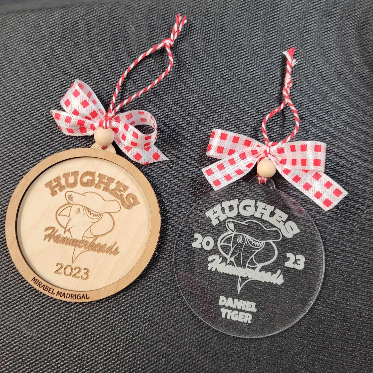 Hughes Hammerheads logo and 2023 on a custom laser engraved holiday and Christmas ornament. The left ornament is made out of maple wood with a red/white bow and string. The right ornament has the design laser engraved on a clear acrylic, both 3.5" in diameter. 